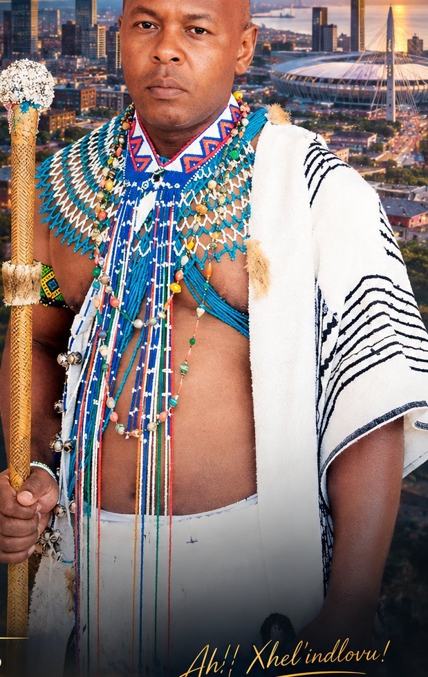 Nkosi Xhel'indlovu in a beaded collar and white blanket, holding a beaded staff in front of the Gqeberha skyline