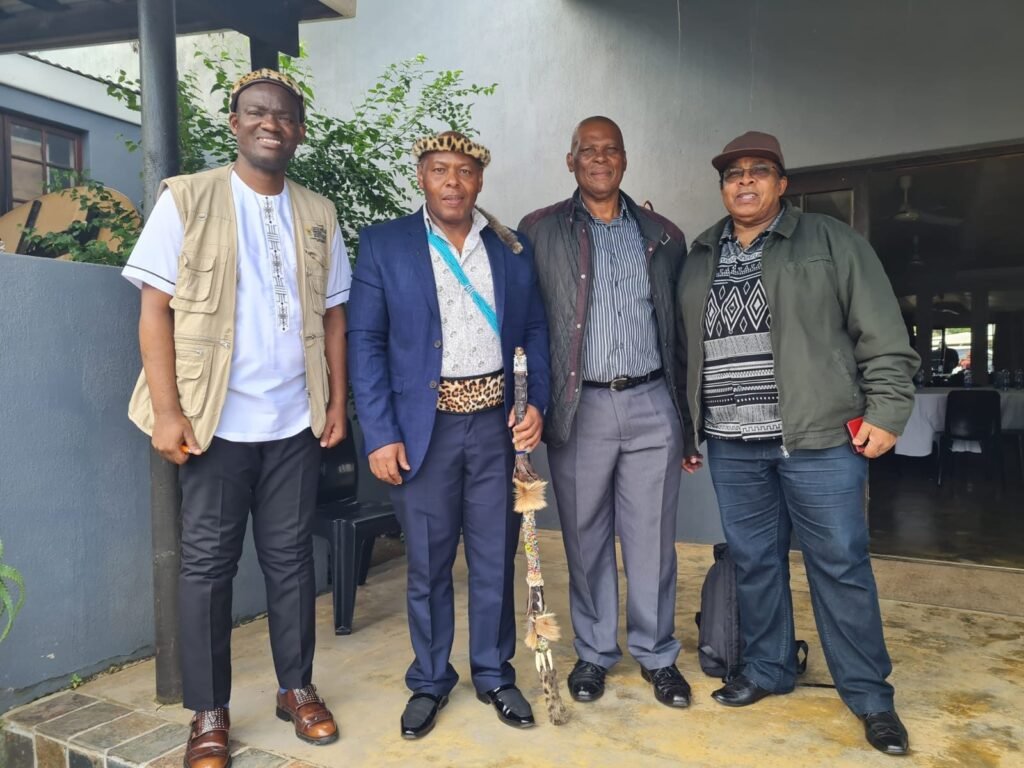 Four men standing together outside a building, one in a leopard-skin hat and beaded sash holding a decorated staff