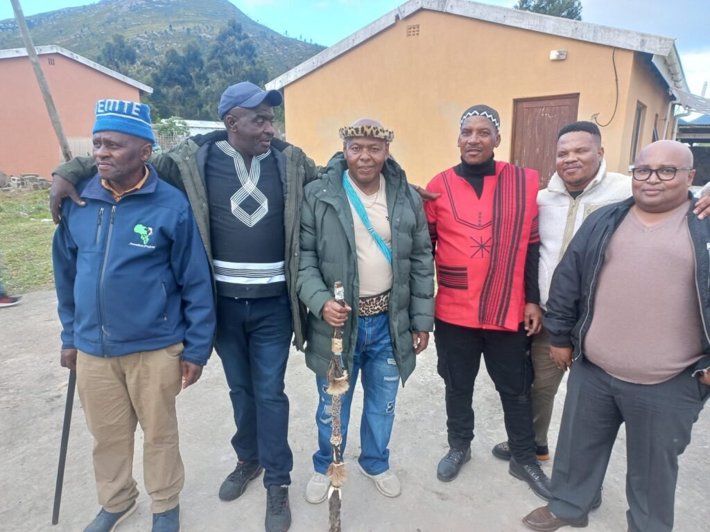 Six men standing together in a village courtyard with a mountain behind, the centre figure holding a beaded staff