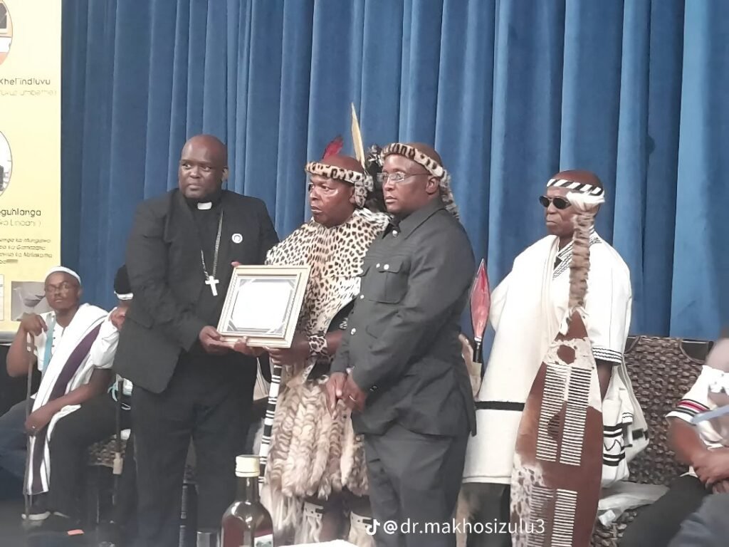 A clergyman presenting a framed certificate to men in leopard-skin regalia on a stage with a blue curtain